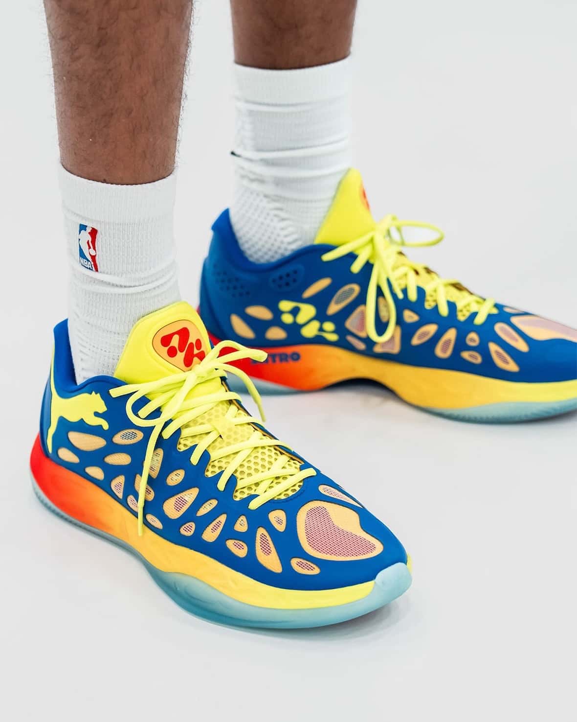 Pies de Tyrese Haliburton con medias blancas y las PUMA Hali 2 Thermal Vision, de capellada azul con aberturas, jaula amarilla flúor, el logo de PUMA en rojo y amarillo y la mediasuela con degradé de rojo a amarillo sobre una suela celeste