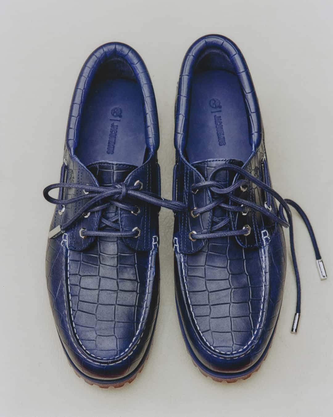 Par del Timberland x Jacquemus Authentic Boat Shoe en navy visto desde arriba sobre fondo gris claro, con todo el cuero en relieve de cocodrilo azul, costuras blancas en la puntera, ojales y herretes plateados, cordones azules y la plantilla azul con el árbol de Timberland y la palabra Jacquemus grabados