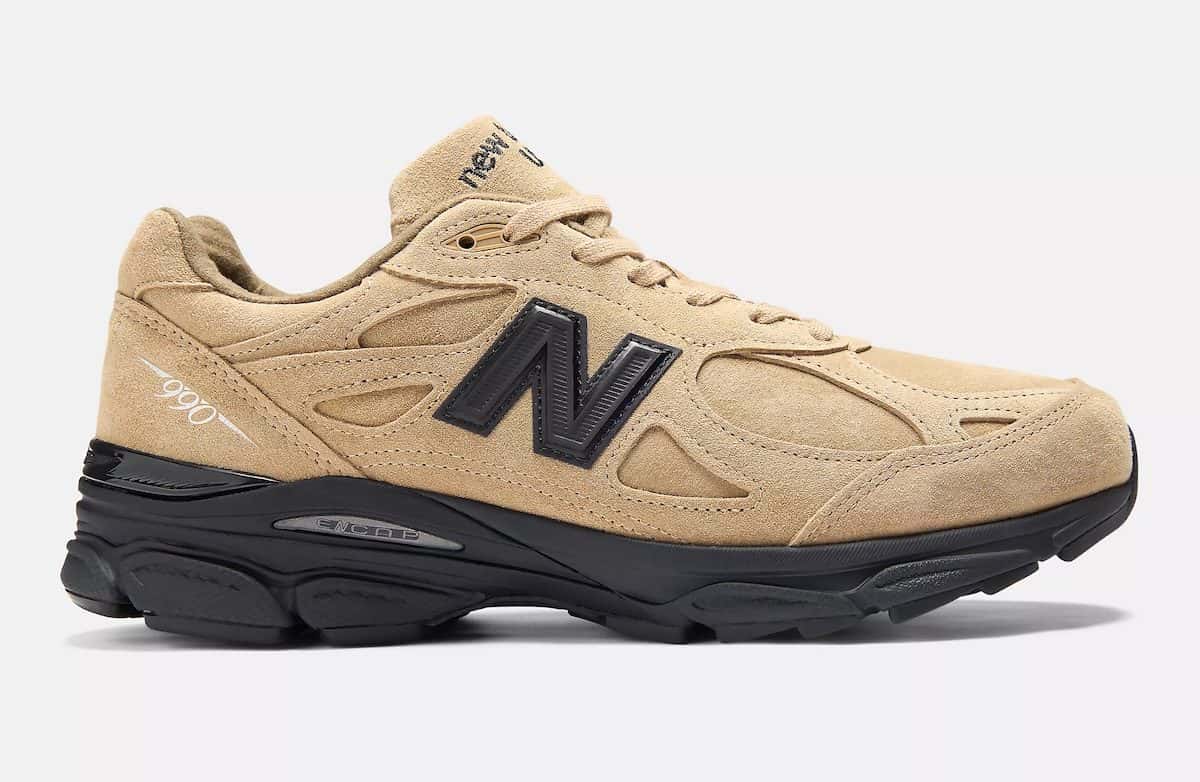 JJJJound x New Balance 990v3 Tan Suede de perfil sobre fondo gris claro, con gamuza tan en todo el upper, la N negra, la leyenda 990 en el talón y la suela negra