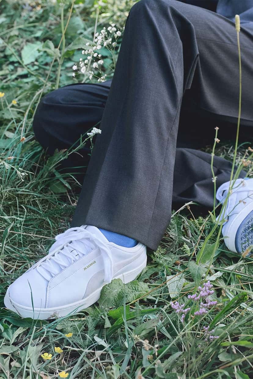 Modelo sentado en el pasto con zapatillas blancas de Jil Sander x PUMA y pantalón de vestir gris