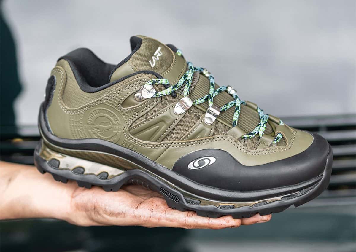 Mano sosteniendo una Salomon XT-Quest 2 de L'Art de L'Automobile en verde oliva, con puntera negra, cordones de soga verdes, ganchos plateados y el logo circular grabado en el talón
