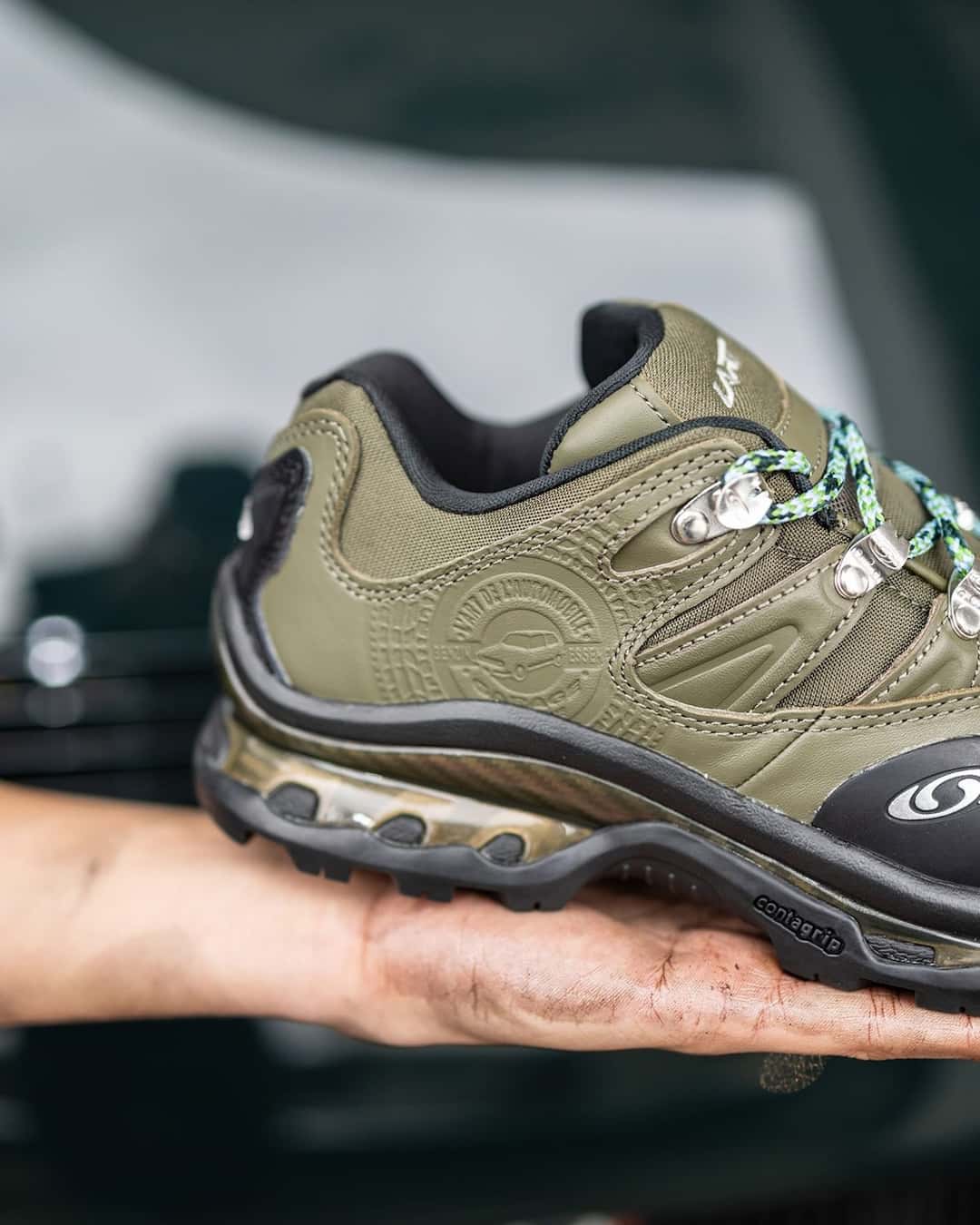 Salomon XT-Quest 2 de L'Art de L'Automobile en verde oliva sostenida con una mano manchada de grasa, con la puntera negra y el logo de Salomon a la vista