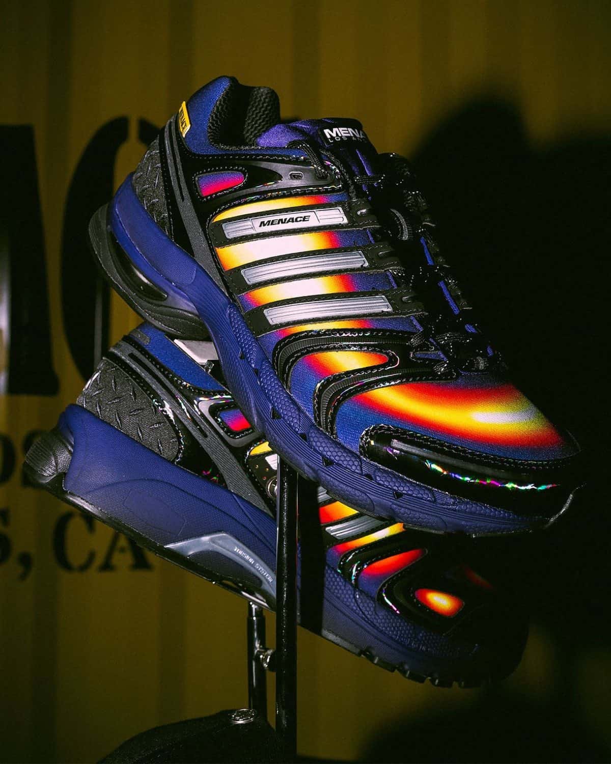 Par de MENACE x adidas Adistar Control 5 Heat Signature sobre un soporte, con degradé amarillo, rojo y blanco tipo cámara térmica, base y mediasuela violeta, overlays negros brillantes y Tres Tiras plateadas con la palabra MENACE