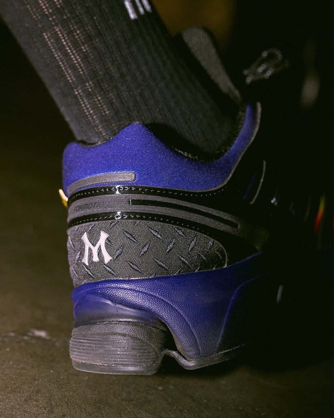 Detalle del talón del MENACE x adidas Adistar Control 5 Heat Signature, con una M blanca sobre un panel negro con textura de chapa antideslizante, cuello violeta y mediasuela violeta
