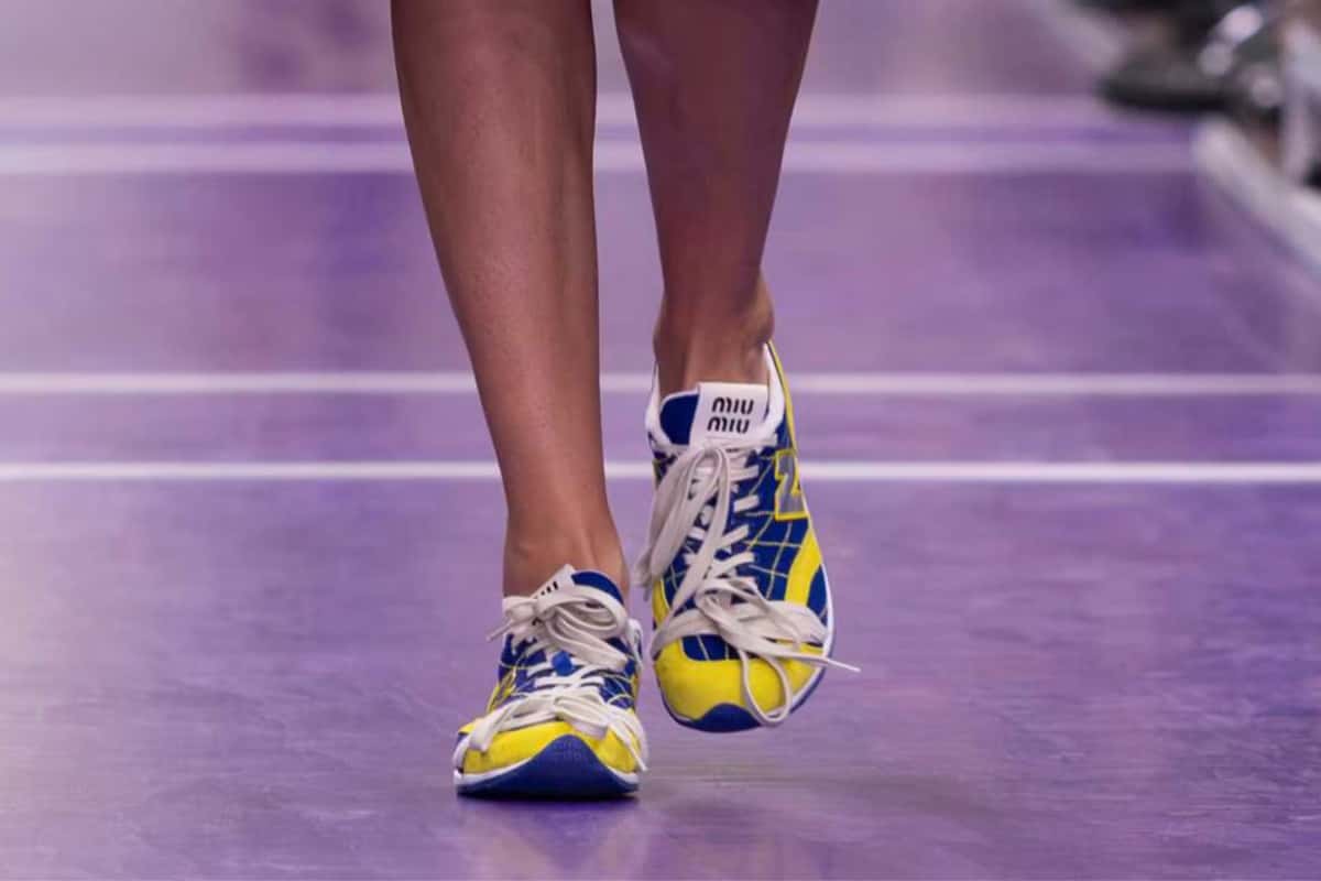 Primer plano de una modelo caminando sobre la pista violeta del desfile Primavera/Verano 2027 de Miu Miu con las New Balance RC150 azules y amarillas, cordones blancos anudados y la etiqueta Miu Miu en la lengüeta