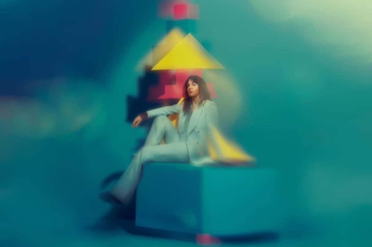 Foto de prensa de Anna Meredith: la compositora, con traje claro, sentada sobre un cubo turquesa frente a figuras geométricas amarillas y rojas desenfocadas, sobre un fondo verde azulado