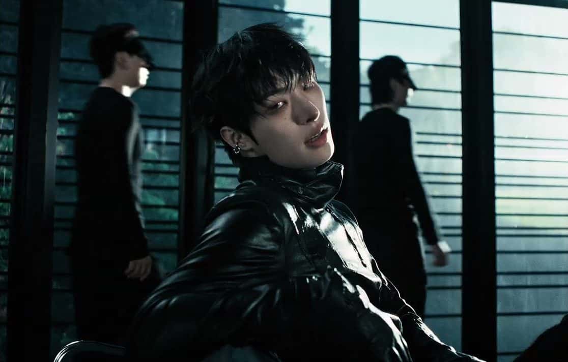 Fotograma del video oficial de «Drip»: un integrante de ATEEZ con campera de cuero negra mira a cámara, con otros dos de fondo frente a un ventanal con rejas