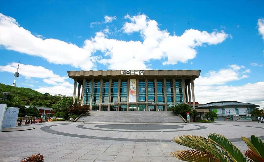 El Teatro Haeoreum del Teatro Nacional de Corea, en Seúl: un edificio de columnas de hormigón y paredes vidriadas con el nombre en coreano arriba, al final de una escalinata y una plaza, con la torre N Seoul de fondo a la izquierda. Foto: Korea.net / Korean Culture and Information Service, Wikimedia Commons, CC BY-SA 2.0