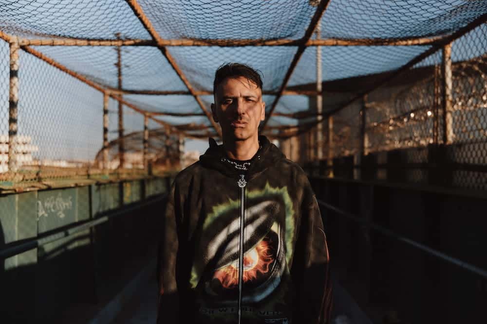 Boys Noize con buzo negro estampado, parado de frente en una pasarela cubierta con alambrado y luz de atardecer, en la foto de prensa oficial de la etapa de «7LLVSION»