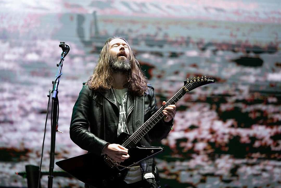 Uno de los guitarristas de la banda de Bruce Dickinson, de pelo largo y barba, toca una guitarra negra frente a una pantalla, en un show de la gira de «The Mandrake Project» en 2024 (foto de archivo). Foto: Dario De Marco, dominio público, vía Wikimedia Commons
