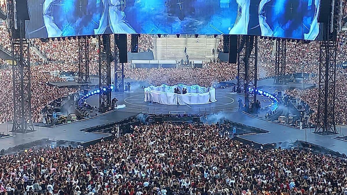 BTS sobre el escenario central de la gira ARIRANG, rodeado de telas blancas y del público en el campo, con pantallas gigantes arriba, en un show en París (foto de archivo, julio de 2026)