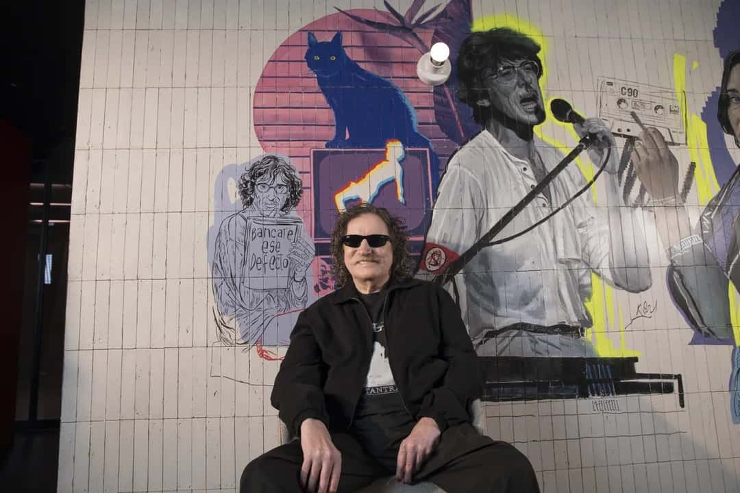 Charly García sentado con anteojos negros frente a un mural pintado con escenas de su carrera, entre ellas un gato azul y una imagen suya cantando, en una imagen del anuncio oficial de Netflix