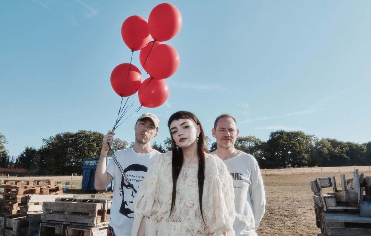 Foto de prensa de Chvrches para «All the King's Men»: Lauren Mayberry, con vestido blanco y pelo largo oscuro, en el centro, entre Iain Cook y Martin Doherty, en un campo con pallets de madera; uno de ellos sostiene un racimo de globos rojos. Foto: Jody Evans