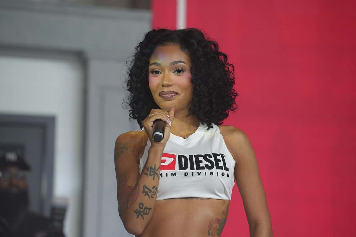 Coi Leray sonríe con el micrófono en la mano sobre el escenario del Essence Festival of Culture, con un top blanco y el pelo rizado, ante un fondo rojo (foto de archivo, julio de 2026)