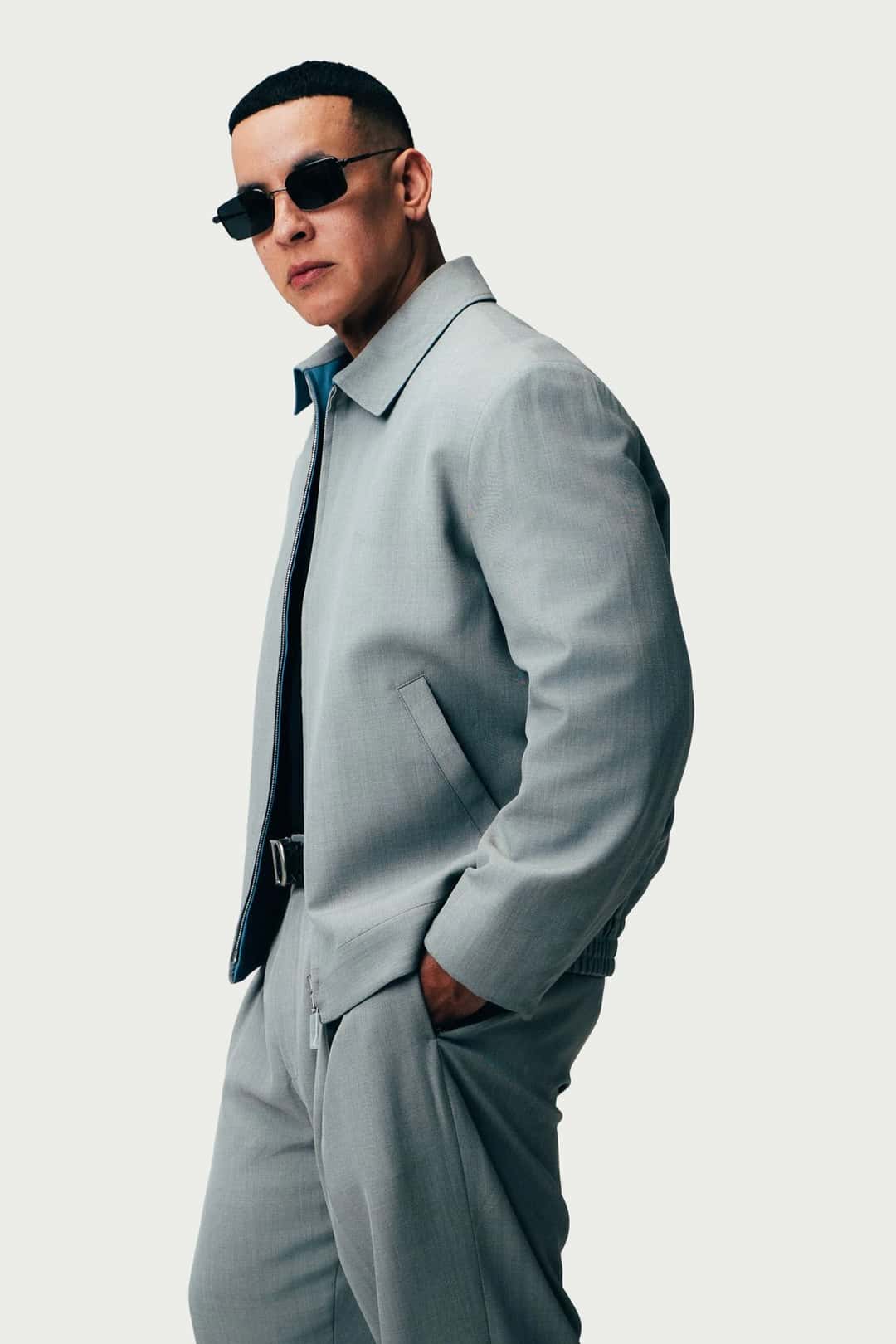 Daddy Yankee de pie, con anteojos de sol, campera y pantalón gris claro y una mano en el bolsillo, en una foto oficial de 2026