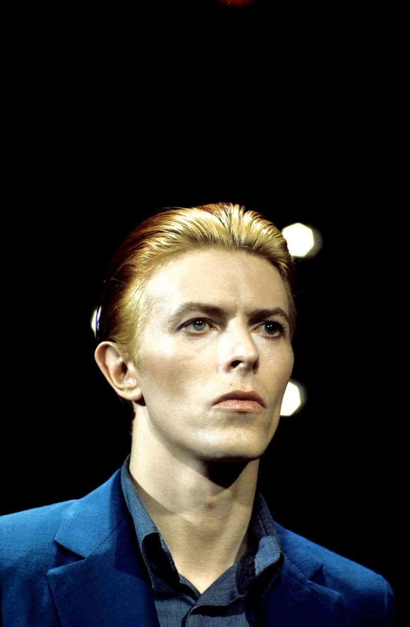 Foto de archivo de David Bowie en los años 70, con el pelo rubio peinado hacia atrás, saco y camisa azules, sobre un fondo negro con luces desenfocadas. Imagen publicada en el sitio oficial de Bowie80