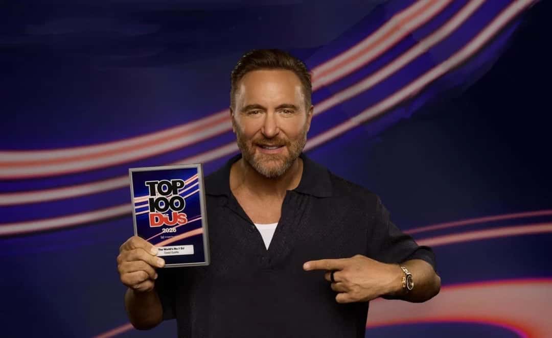 Foto oficial de David Guetta, de camisa negra, sonriendo y señalando con la mano izquierda la placa del Top 100 DJs 2026 que sostiene con la derecha, que dice «The World's No.1 DJ», sobre un fondo azul con líneas de luz rojas