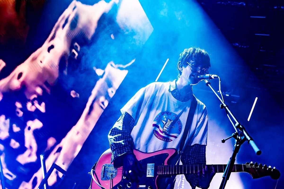 El cantante y guitarrista de DIIV canta y toca una guitarra eléctrica roja frente al micrófono, con remera blanca y anteojos, bajo luces azules, en septiembre de 2025 (foto de archivo)