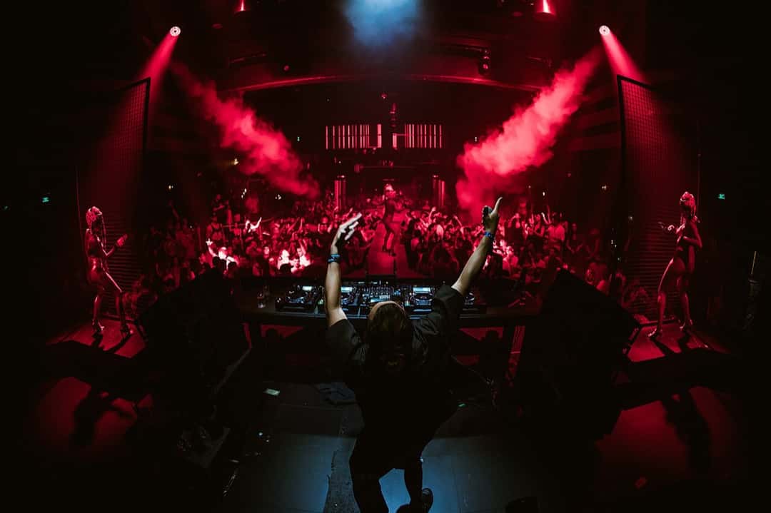 Un DJ de espaldas con los brazos en alto frente a la cabina de Hï Ibiza, con la pista llena, humo rojo y bailarinas a los costados del escenario. Foto: Simone Martellarci/Marte, Wikimedia Commons, CC BY 3.0