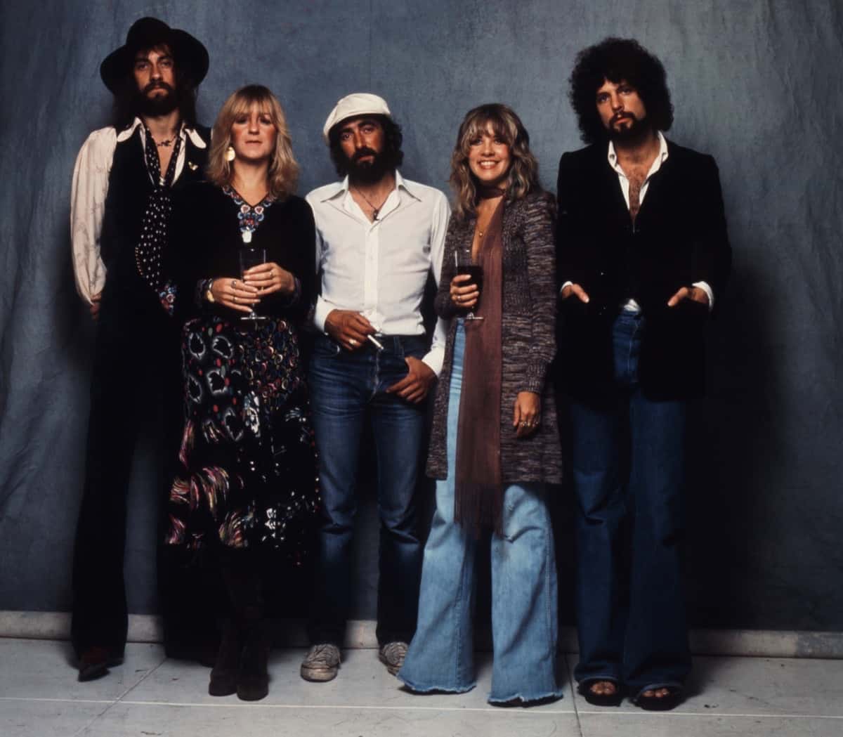 Foto oficial del anuncio de «Fleetwood Mac: The Chain»: Mick Fleetwood, Christine McVie, John McVie, Stevie Nicks y Lindsey Buckingham posan de pie en los años setenta frente a un telón gris azulado, con ropa de época y copas en la mano