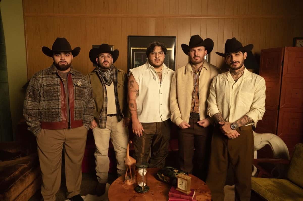 Foto de prensa de los cinco integrantes de Grupo Frontera parados en un living con paredes de madera, cuatro con sombrero texano negro, frente a una mesa ratona con un reloj de arena y un reloj de mesa