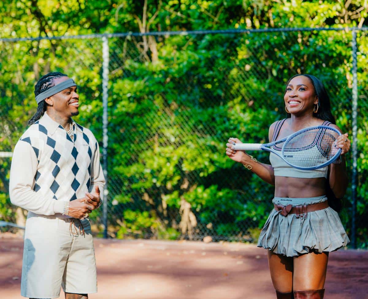 Foto oficial del rodaje del video de "Pop It": Gunna, con vincha gris y un buzo con rombos, se ríe junto a Coco Gauff, que sostiene una raqueta en una cancha de tenis rodeada de árboles