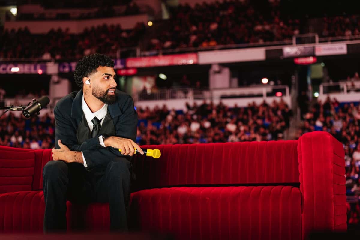 Foto de prensa de Jay Wheeler, de traje oscuro y corbata negra, sentado en un sillón de terciopelo rojo con un micrófono amarillo en la mano, frente a las tribunas llenas de un estadio cubierto