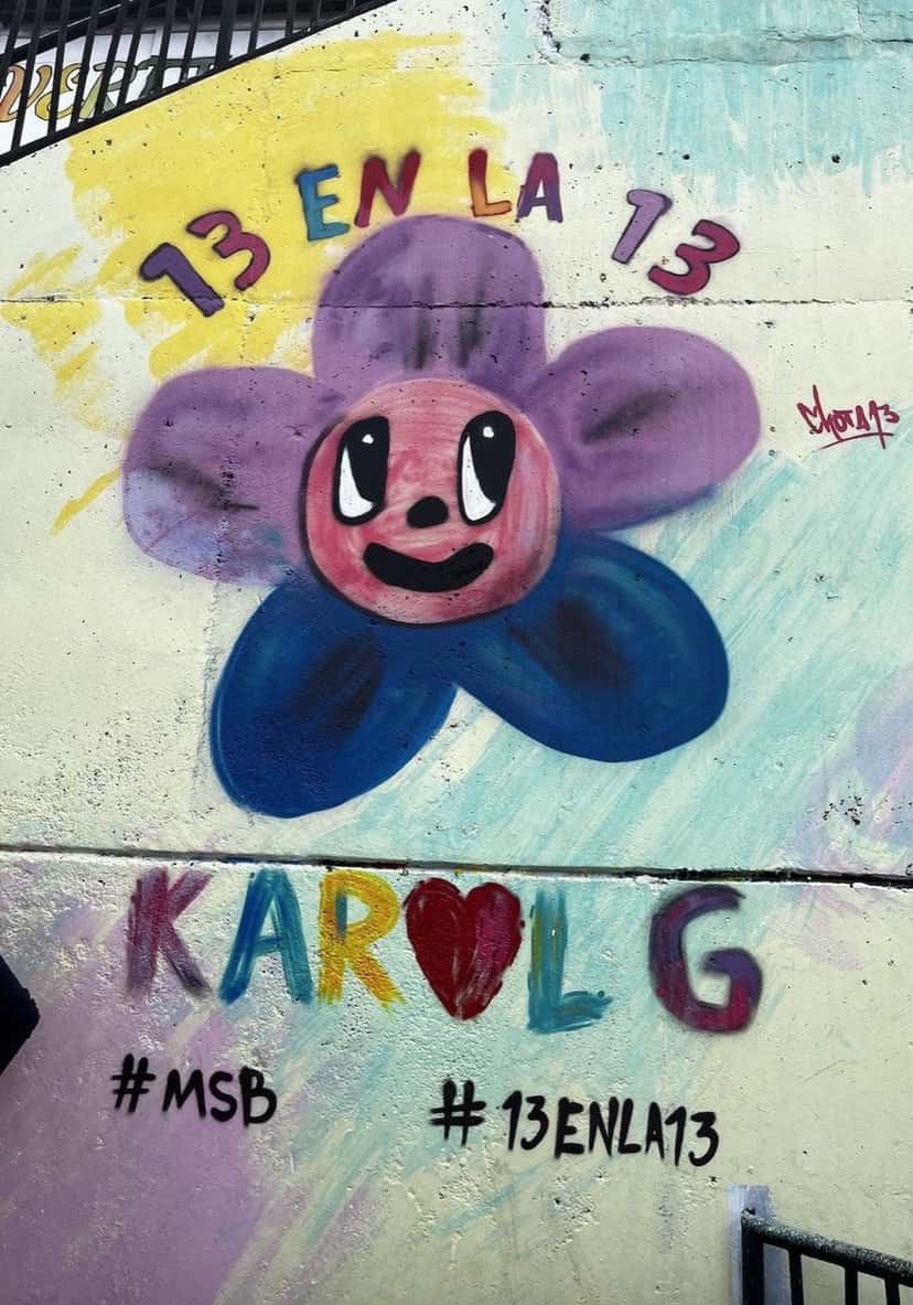 Mural de Karol G en la Comuna 13 de Medellín, en 2023: una flor violeta y azul con cara sonriente, la frase «13 en la 13», el nombre KAROL G con un corazón rojo en lugar de la O y los hashtags #MSB y #13enla13 (foto de archivo). Foto: Alecprofit, CC BY-SA 4.0, vía Wikimedia Commons