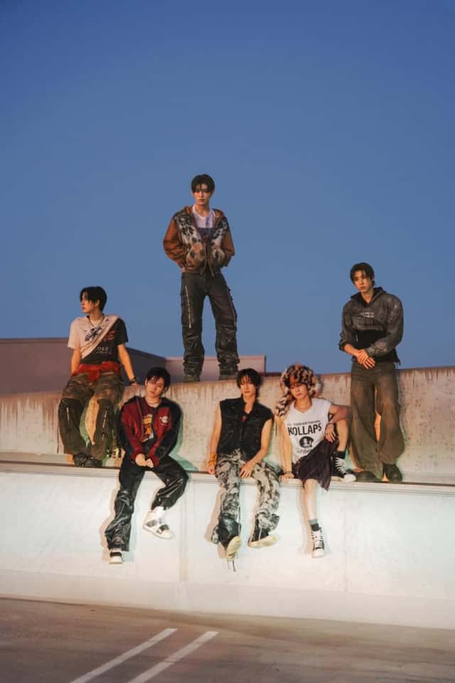 Foto de prensa oficial de JYP Entertainment de KickFlip: seis integrantes posan al atardecer sobre un muro de cemento en una terraza, tres sentados en el borde y tres parados, con una campera bomber bordó, chalecos y camperas de cuero, pantalones cargo y camuflados, zapatillas altas y un gorro de piel con estampado animal, bajo un cielo azul