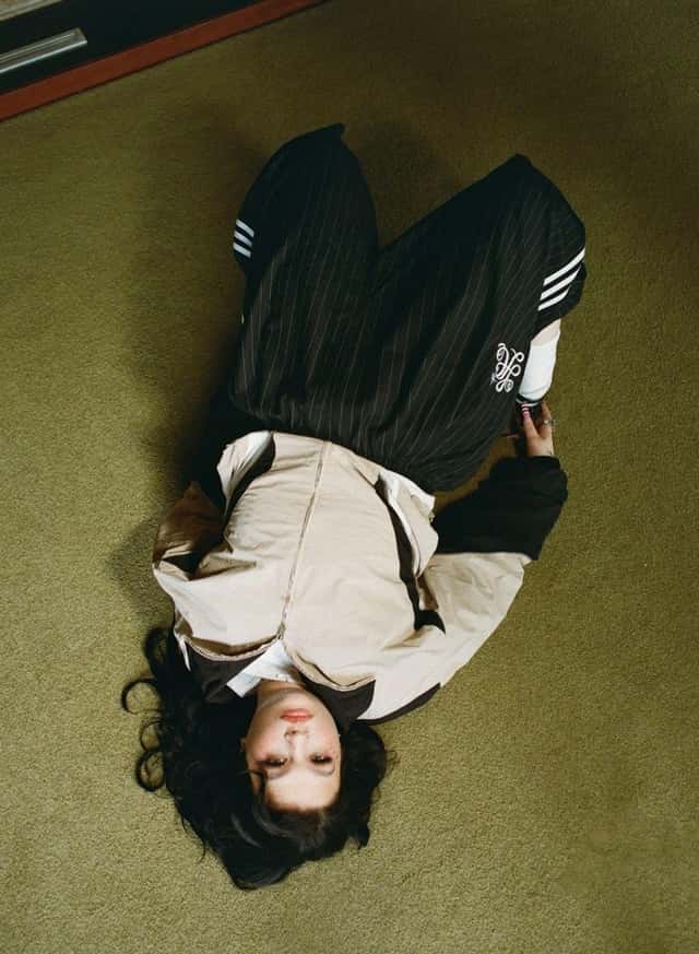 Foto de prensa de Lola Young acostada boca arriba sobre una alfombra verde, vista desde arriba, con campera beige y negra y pantalón negro a rayas finas