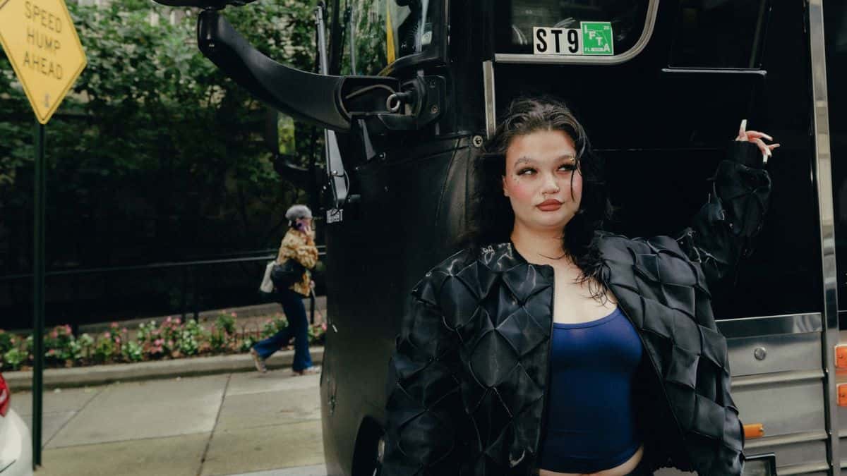 Lola Young con campera negra de cuero acolchada y top azul, apoyada contra un micro negro en una vereda arbolada, con un cartel amarillo de tránsito a la izquierda. Foto: Harriet K Bols