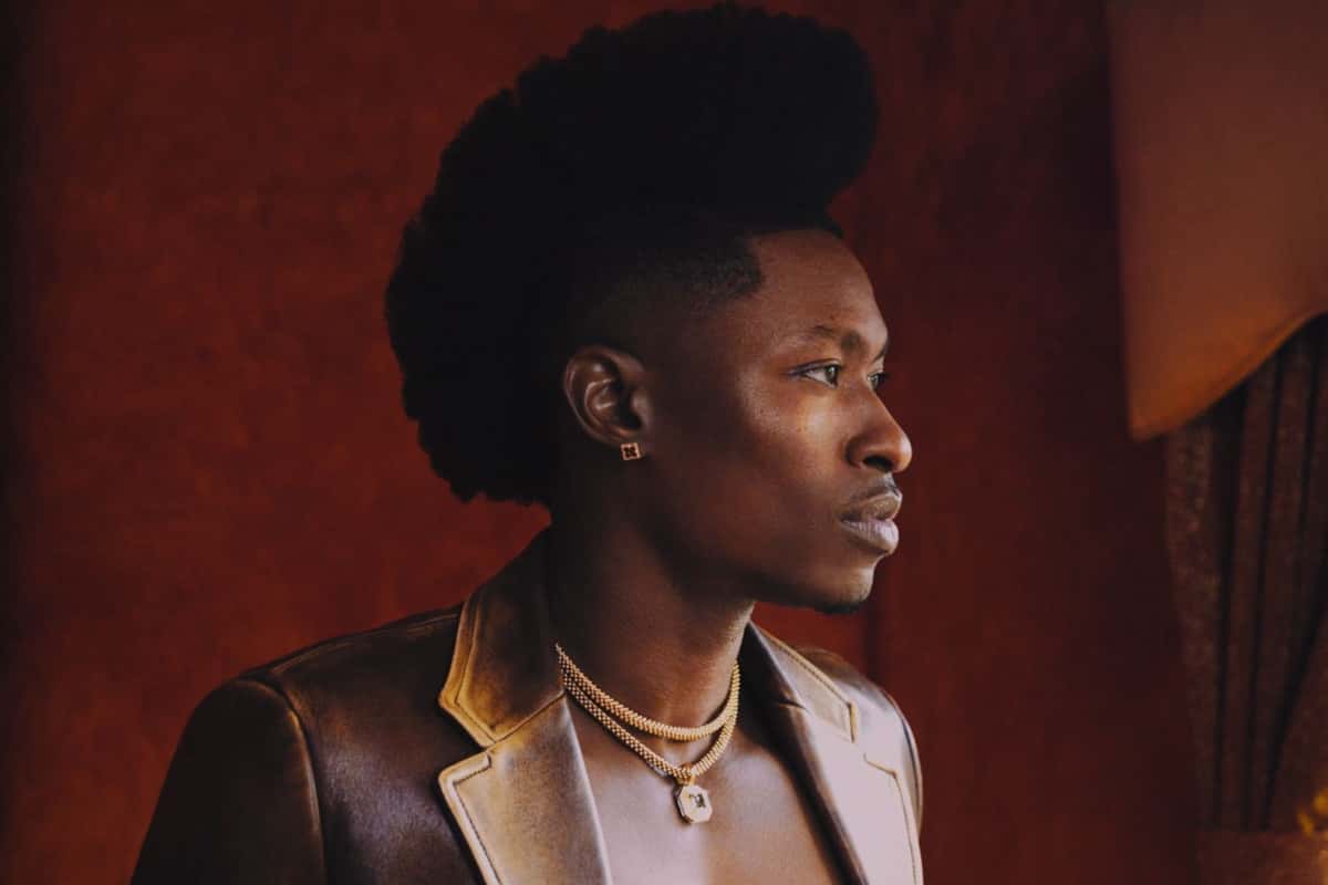 Retrato de prensa de Lucky Daye de perfil, con saco de cuero marrón, collares dorados y el pelo alto, frente a una pared bordó. Foto: Micaiah Carter