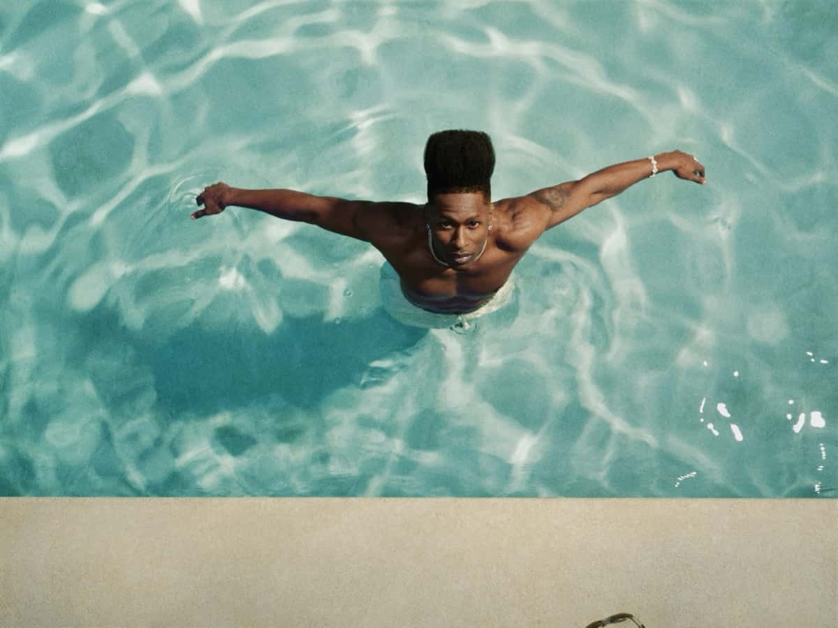 Foto de prensa oficial de Lucky Daye, vista desde arriba: el cantante, con el pelo alto, cadena plateada y pulsera, está metido en una pileta de agua turquesa con los brazos abiertos y mira a cámara, junto al borde de piedra clara. Foto: Micaiah Carter, cortesía de Warner Records