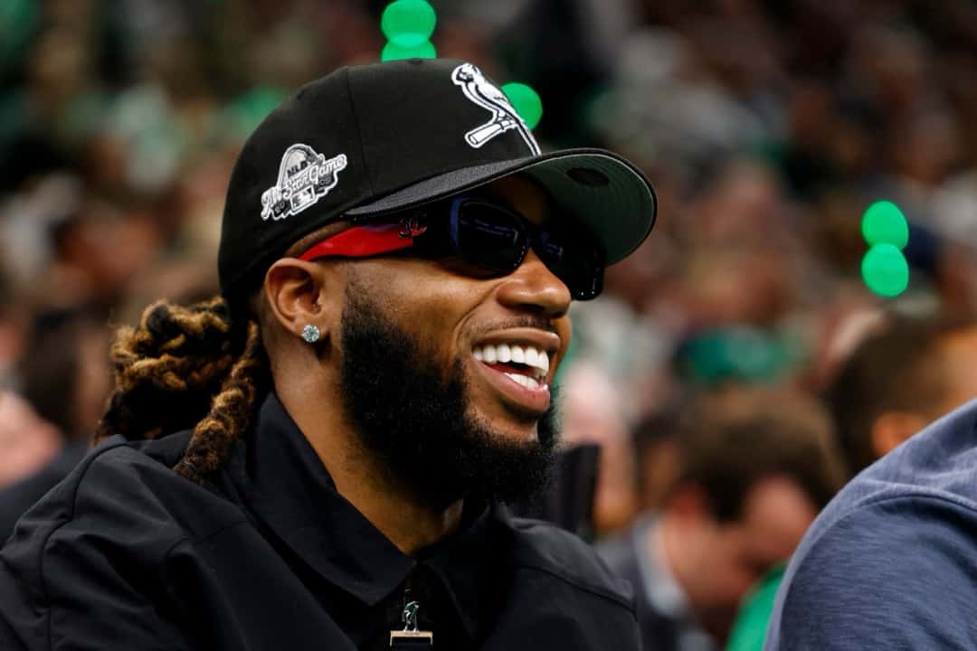 Metro Boomin sonriendo en la tribuna de un partido de básquet, con gorra negra, anteojos oscuros de marco rojo y camisa negra