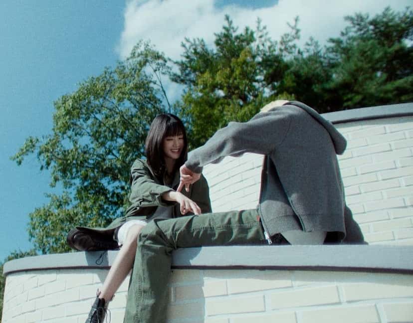 Fotograma del teaser de «to the moon :)»: MINNIE, con campera verde, sentada sobre un muro blanco junto a una persona de buzo gris, con árboles y cielo de fondo