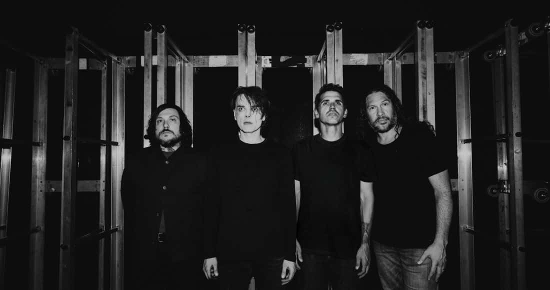 Foto de prensa oficial de My Chemical Romance en blanco y negro: los cuatro integrantes, vestidos de negro, posan de pie y serios entre estructuras metálicas de escenario, sobre un fondo oscuro