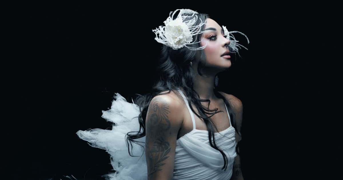 Pabllo Vittar de perfil sobre fondo negro, con pelo largo oscuro, tocado de flor blanca con plumas y vestido blanco de tul