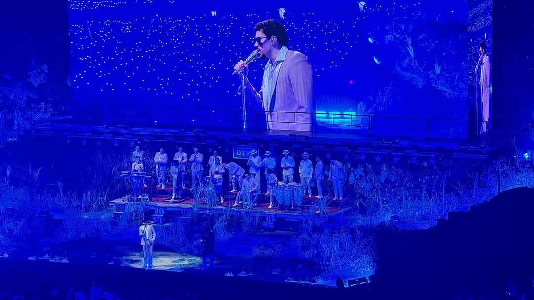 Foto de archivo de Bad Bunny en vivo en septiembre de 2025: canta con micrófono y saco claro en una pantalla gigante, mientras abajo, en un escenario con plantas y bajo luz azul, lo acompaña una banda vestida de blanco. Foto: Comecoquito, Wikimedia Commons, CC0