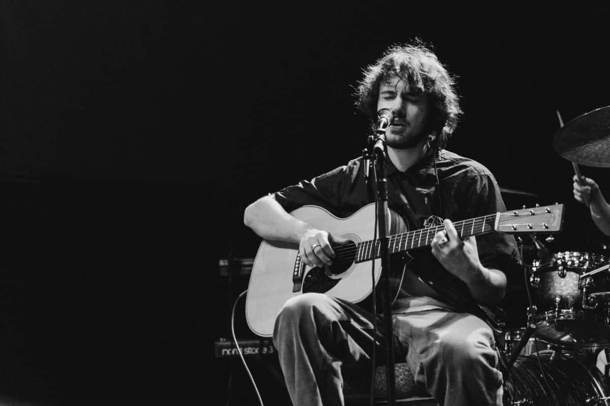 Foto de archivo en blanco y negro de Quadeca sentado en el escenario, cantando con los ojos cerrados frente al micrófono mientras toca una guitarra acústica, con la batería detrás