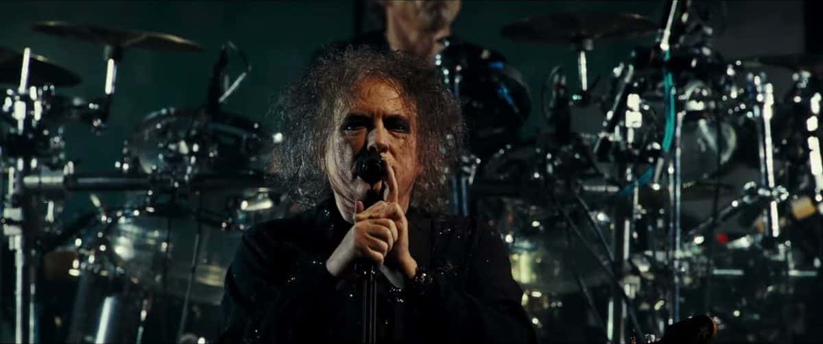 Fotograma del video oficial de The Cure en el Troxy de Londres en 2024: Robert Smith, con el pelo negro despeinado, canta tomando el micrófono con las dos manos, frente a la batería (foto de archivo)
