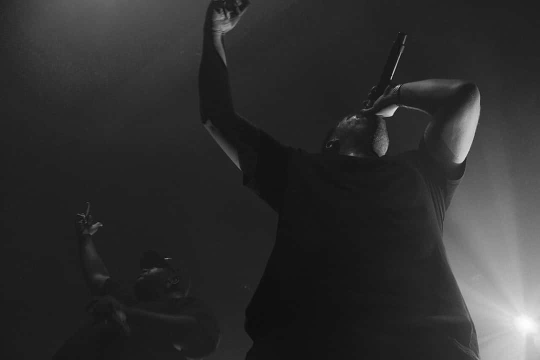 Run the Jewels en vivo, en blanco y negro: uno de los raperos canta con el micrófono en una mano y el otro brazo levantado, con su compañero detrás, en octubre de 2015 (foto de archivo). Foto: Julio Enriquez, CC BY 2.0, vía Wikimedia Commons