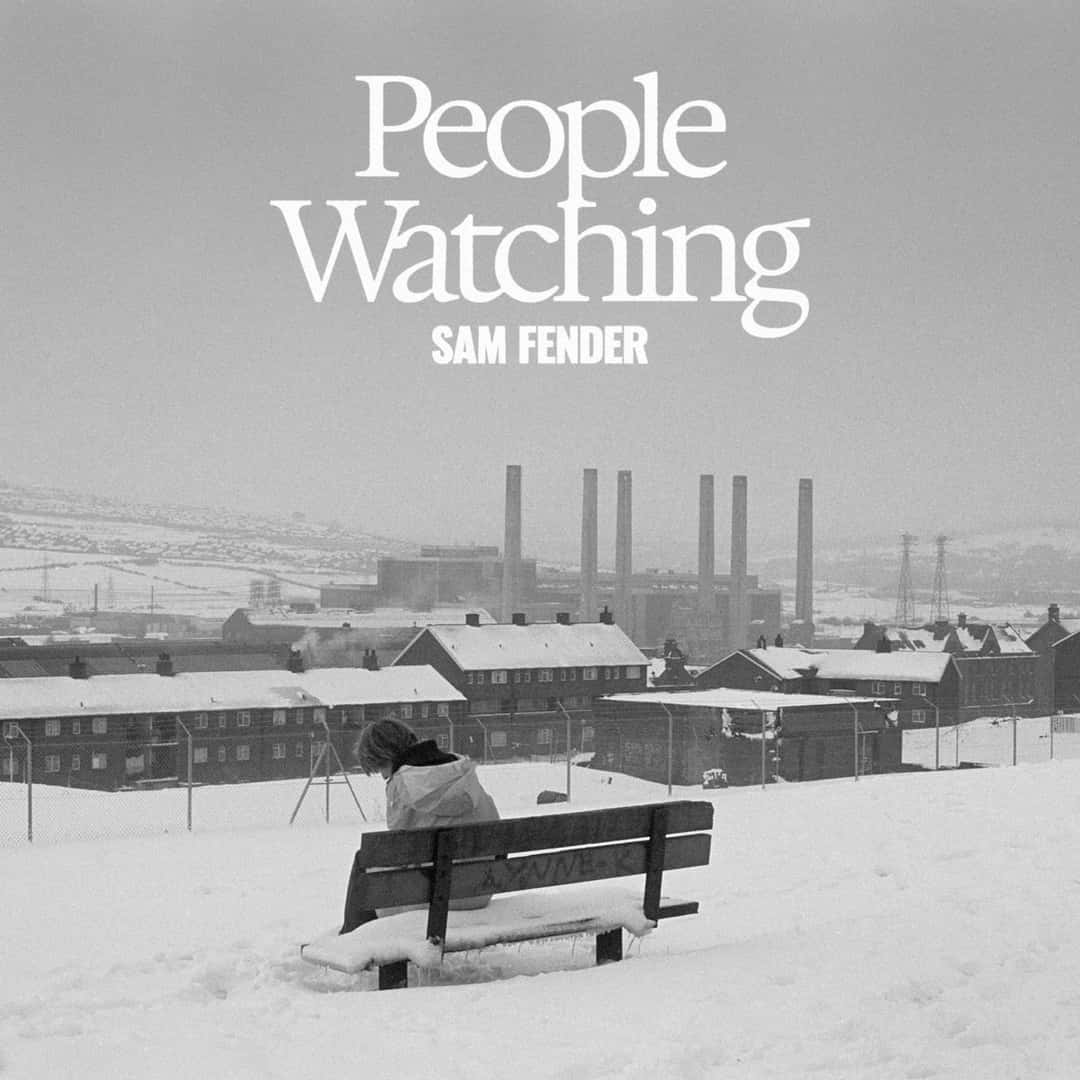 Portada de «People Watching», de Sam Fender: una persona sentada en un banco cubierto de nieve frente a un barrio con chimeneas industriales, en blanco y negro