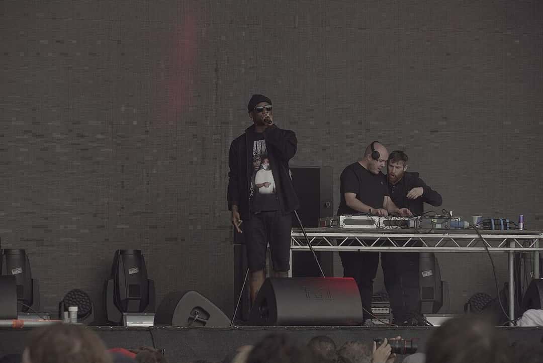 Foto de archivo de Skepta en el escenario principal del festival Field Day de Londres, en junio de 2016: canta con el micrófono en la mano, de negro y con gorra, junto a una mesa de DJ con dos personas detrás. Foto: Jwslubbock, Wikimedia Commons, CC BY-SA 4.0
