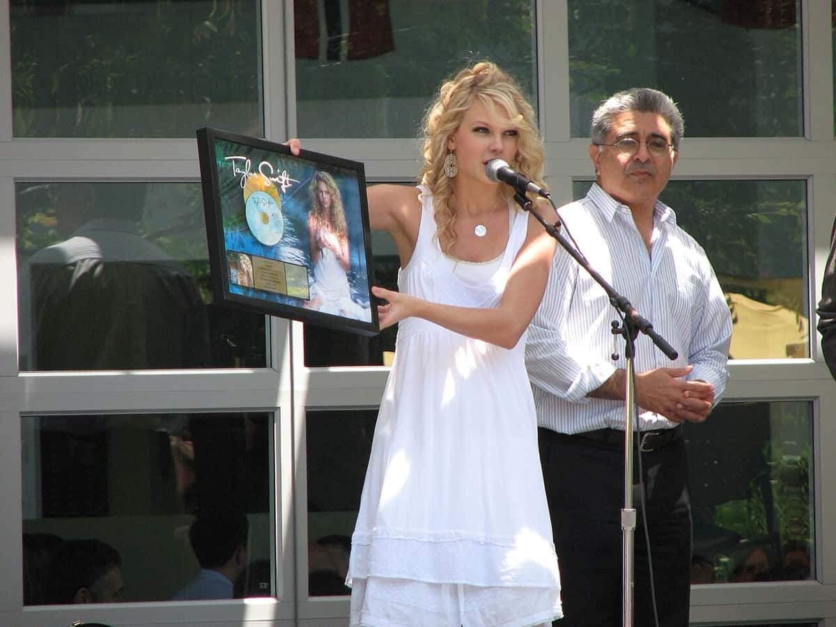 Taylor Swift, de vestido blanco y pelo rubio con rulos, muestra una placa enmarcada de su primer disco frente a un micrófono, en 2007 (foto de archivo)