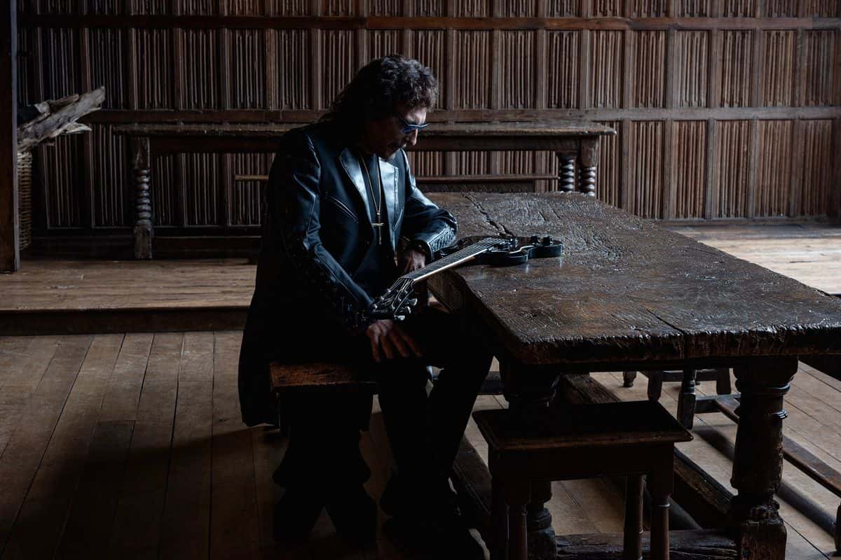 Foto de prensa oficial de Tony Iommi para «From the Dark»: con campera de cuero negra y anteojos, sentado a una mesa larga de madera en un salón antiguo con paredes de madera, sostiene una guitarra eléctrica