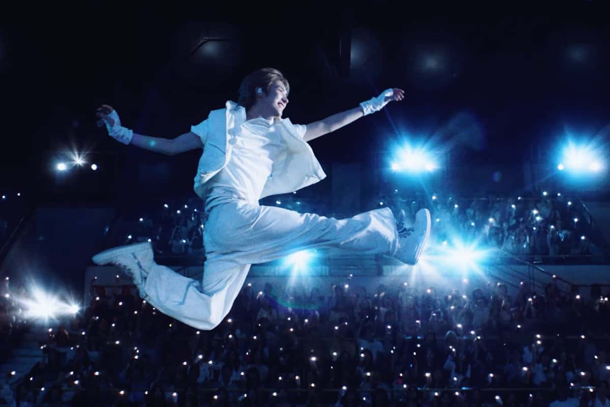 Imagen oficial del tráiler "Being Together : TO US": un integrante de TWS, vestido de blanco, salta con las piernas abiertas sobre un estadio lleno de luces de celulares