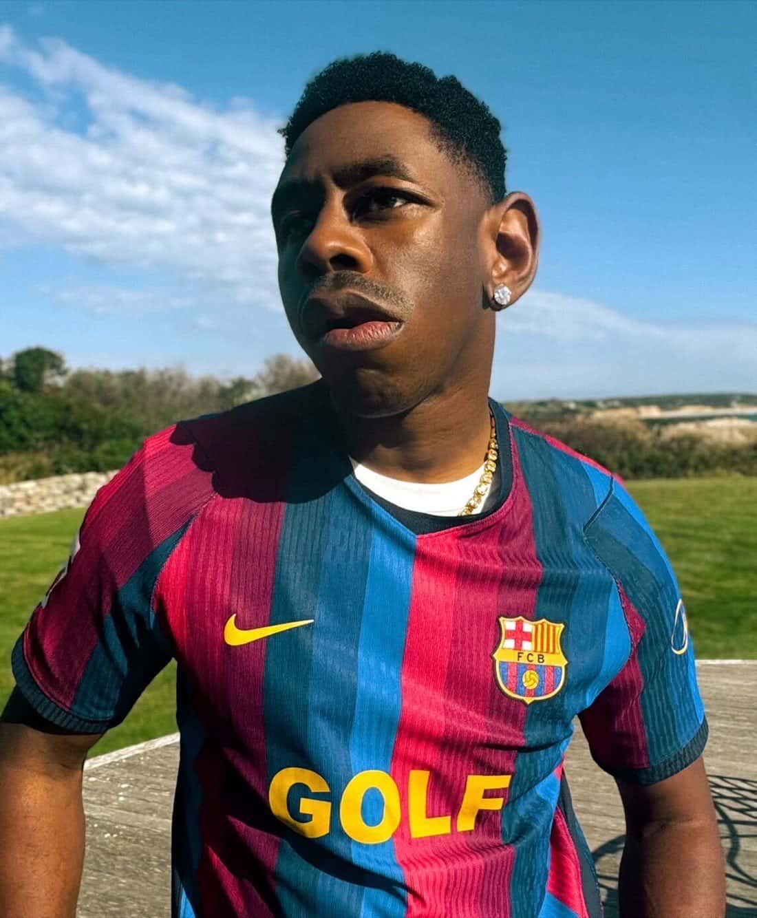 Tyler, The Creator posa al aire libre con la camiseta azul y grana del Barcelona que lleva el logo amarillo de GOLF en el pecho, en una foto de prensa de la colaboración