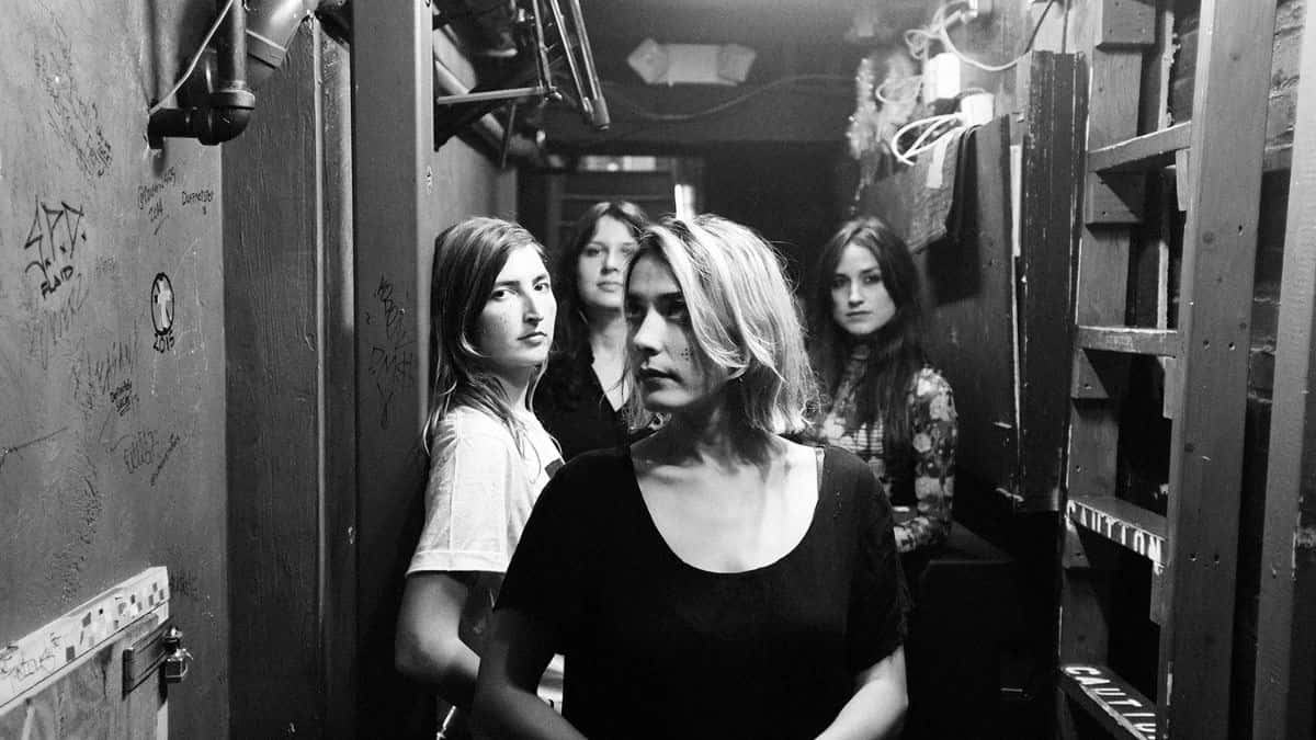 Foto de prensa en blanco y negro de las cuatro integrantes de Warpaint en un pasillo de camarines angosto, con paredes escritas