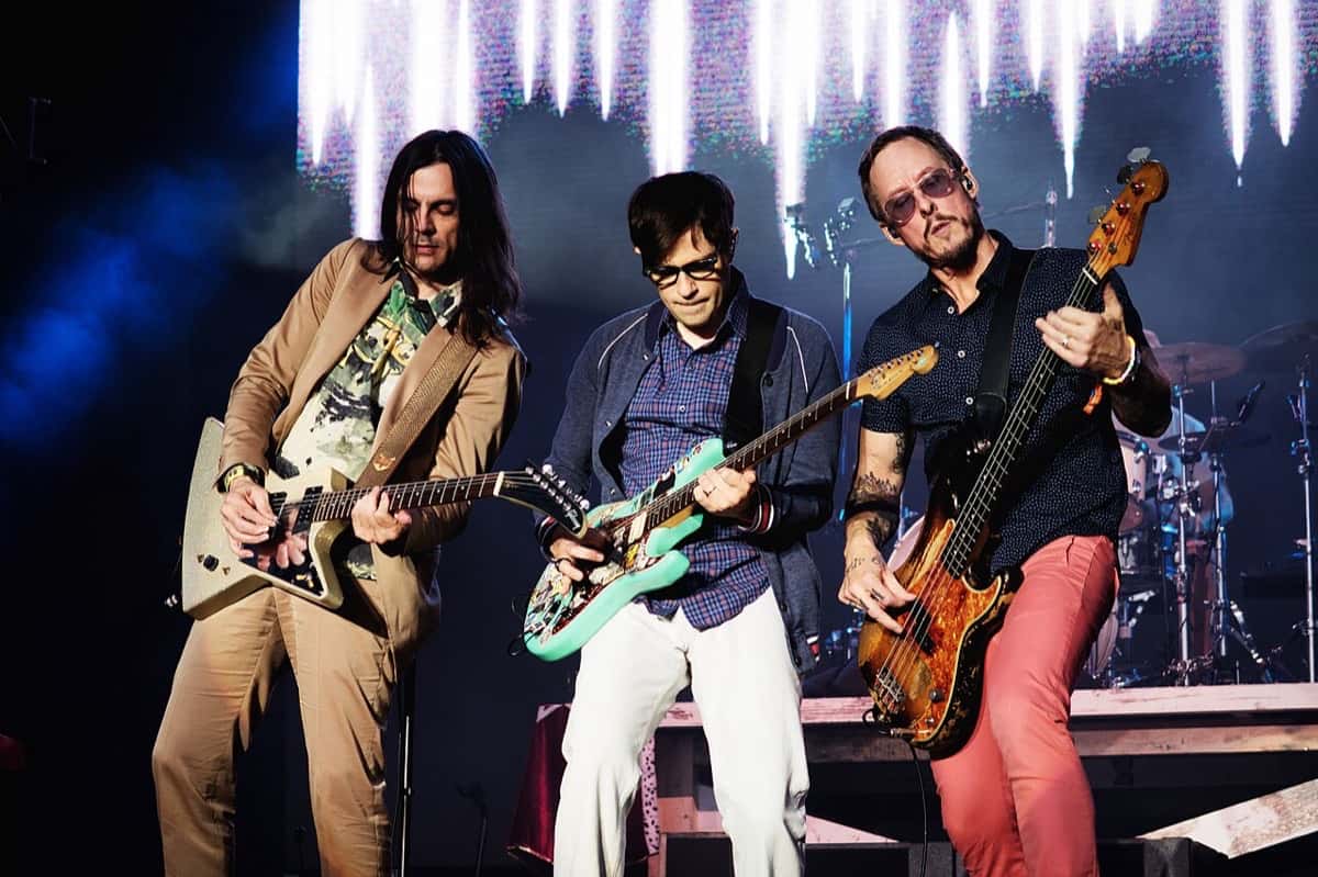 Brian Bell, Rivers Cuomo y Scott Shriner, de Weezer, tocan juntos sus guitarras y el bajo en el escenario, frente a una pantalla con luces blancas (foto de archivo, septiembre de 2017)