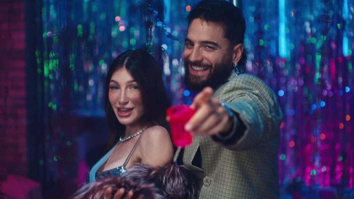 Yami Safdie, de pelo largo castaño y vestido celeste con brillos, y Maluma, con saco a cuadros, sonríen en una fiesta con cortinas de luces de colores mientras él levanta un vaso rosa, en una imagen del video oficial de «Felicidades»